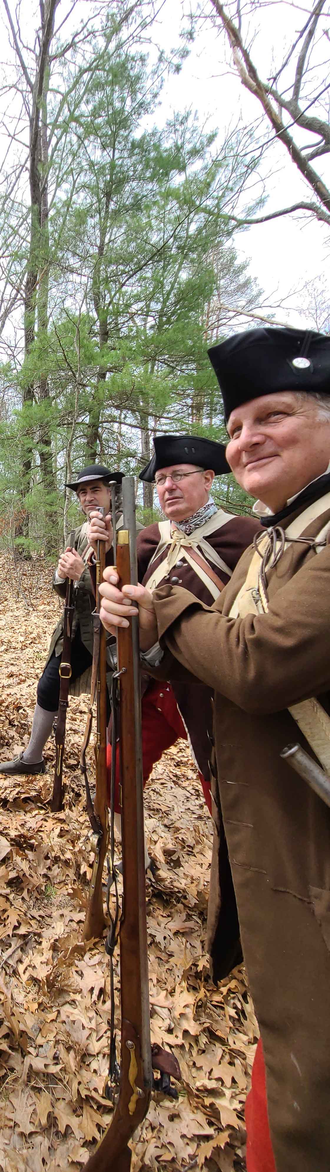 Mike-Sutila,-Bob-Stiles,-and-Mike-Audette-of-the-Acton-Minute-Men-wait-in-the-woods-for-the-arrival-of-the-British-column-at-Battle-Road-2023.-Photo-by-Steve-Crosby.jpg